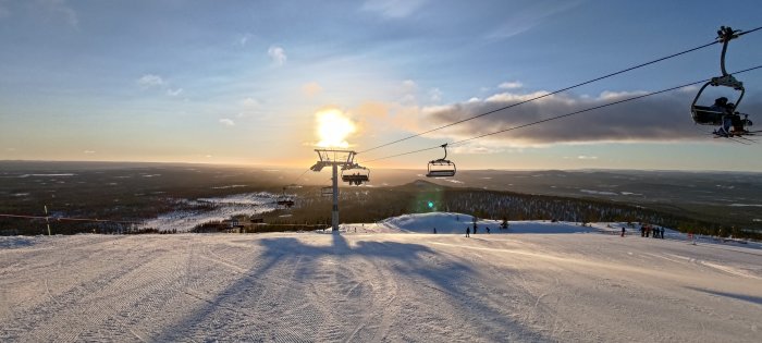 Solnedgång över snötäckt skidbacke med skidlift och skidåkare i vinterlandskap.