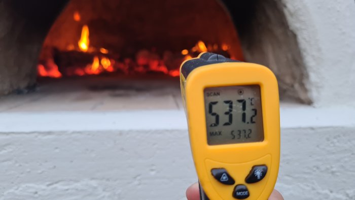 Infraröd termometer visar 537.2°C framför en brinnande eldstad eller ugn.