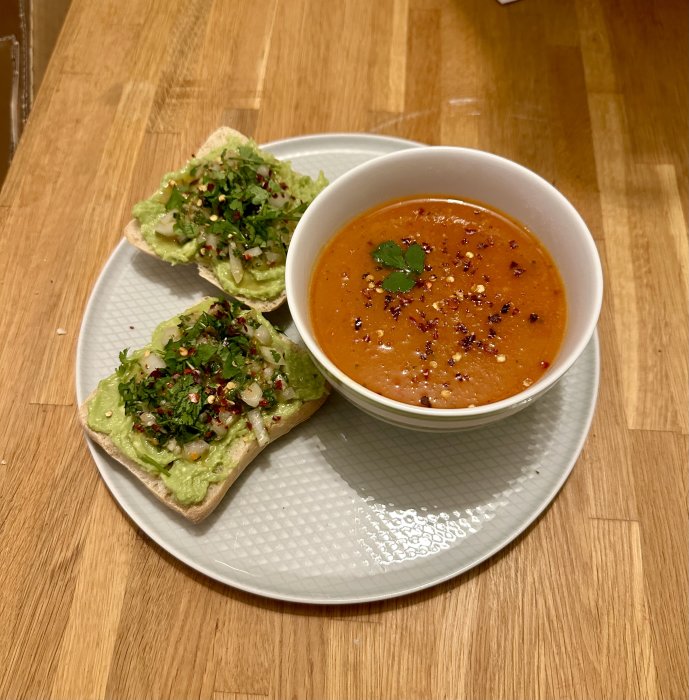 Tomatsoppa i vit skål, avokadomacka med kryddor, träbord, hälsosam lunch, välkryddad, matfotografi.