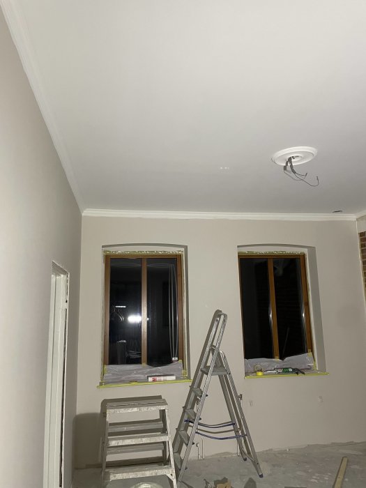 Renoveringsarbete pågår inne i rum med stege, oskyddade fönster och osatt taklampa.