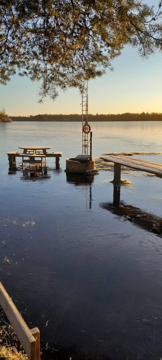En isbelagd brygga med en picknickbord och simstegar i solnedgången, omgiven av stilla vatten och träd.