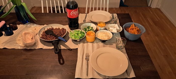 Middagsbord med taco-tillbehör, dryck, tallrikar och bestick förberett för måltid.