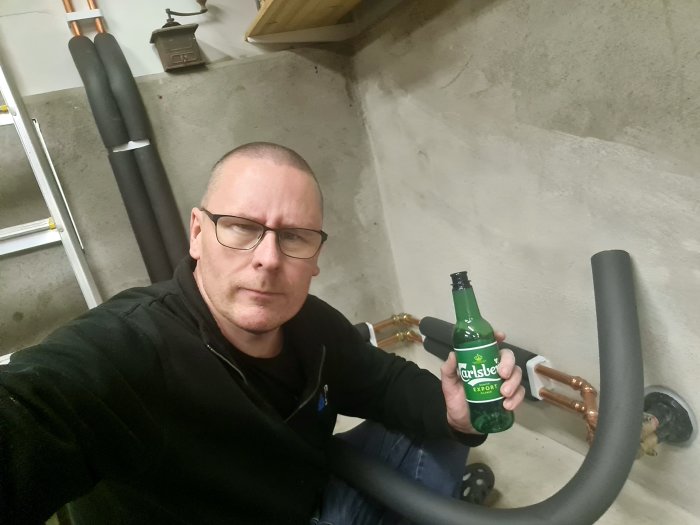 Man tar selfie, håller i ölflaska, sitter på golvet, rörledningar i bakgrunden, källarutrymme.