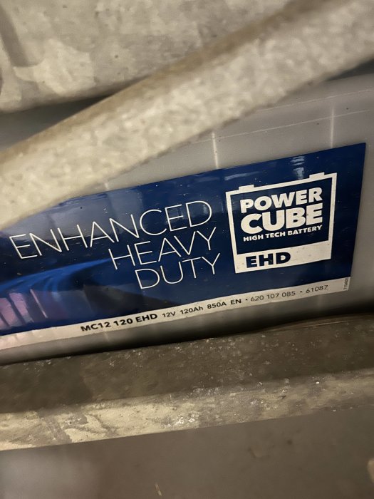Etikett för 'Power Cube Enhanced Heavy Duty'-batteri, mörk bakgrund, dold delvis av struktur.