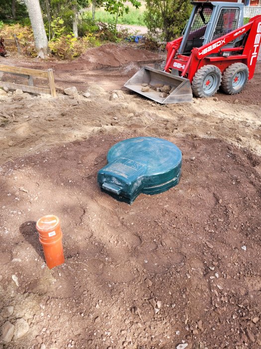 Markarbetsplats med skidsteer, grön septiktanklock, orange rör i skogsmiljö.