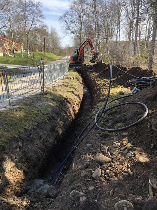 Grävmaskin vid grävd ränna, kablar på marken, bostadsområde i bakgrunden, gathinder, träd och soligt väder.