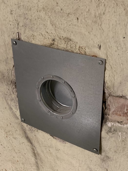 Svart ventilationsspjäll på betongvägg med synliga skruvar och en tegelsten.