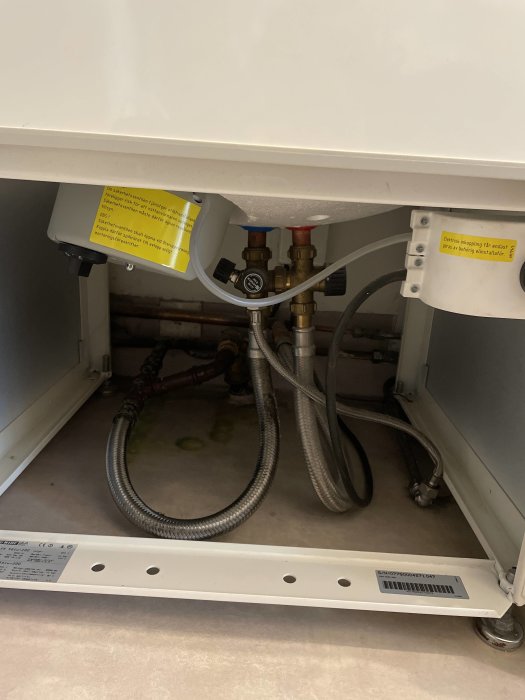 Installationsutrymme under vit skåp med slangar, rör, ventil och varningsetiketter synliga.