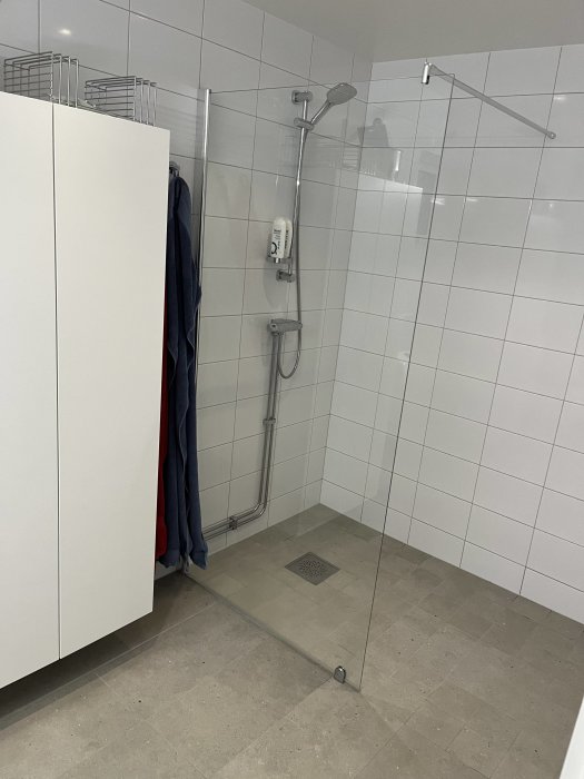Modernt badrum, duschområde, glaspartition, vita kakelväggar, gråa klinkergolv, skåp och handdukshängare.