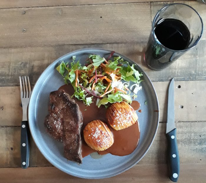 En måltid med stek, hasselbackspotatisar, sallad och ett glas mörk dryck, serverad på ett träbord.