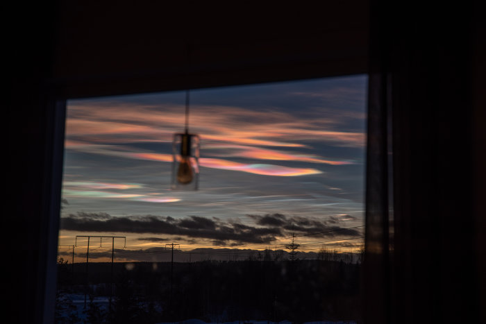 Genom fönster: storslagen solnedgång med färgsprakande himmel och silhuett av landskap. Hängande lampa i förgrunden.