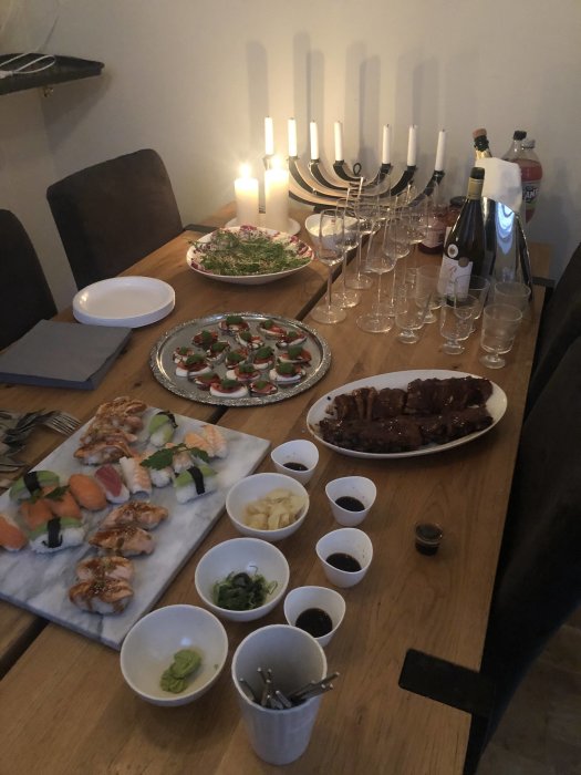 Middagsbord med sushi, ribs, aptitretare, sallad, ljus, dricksglas, och en flaska vin.