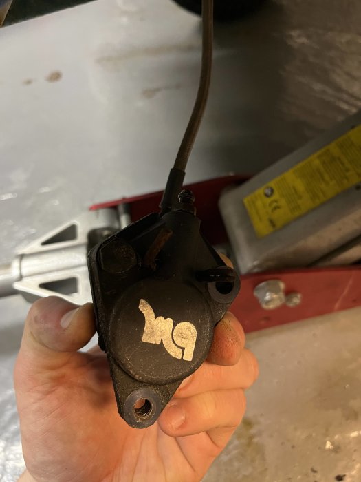 Hand håller rostigt bilkomponent, troligen en bromsok, kabel ansluten, verktyg och vagga i bakgrunden.