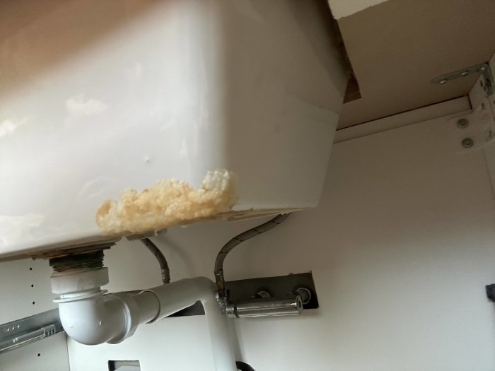 Handfat med vattenlås och rör, underifrån perspektiv, skadat trä och möjlig fuktskada synlig.