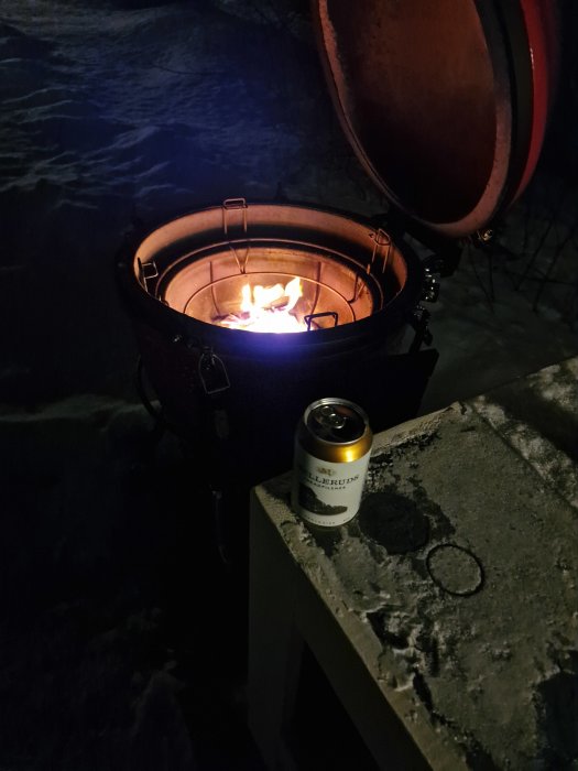 Eld i grill utomhus på vintern, ölburk på betongbänk. Natt, mörkt, kallt, snöigt.