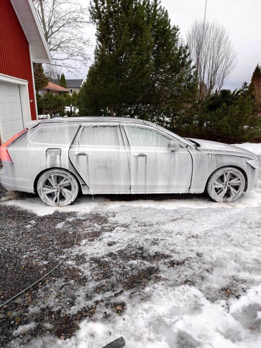 Bil täckt med skum, står vid ett hus, snö, ivasketablering eller desinficering, gråmulet väder.