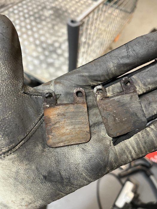 Använd handskar som håller i två slitna bromsbelägg från ett fordon. Arbetsmiljö i bakgrunden.