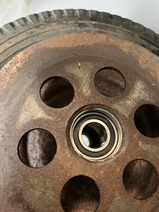 Rostig mekanisk del, försänkta hål, någon form av kugghjul eller remhjul, central lagerinfattning. Metallisk konstruktion, industriell komponent.