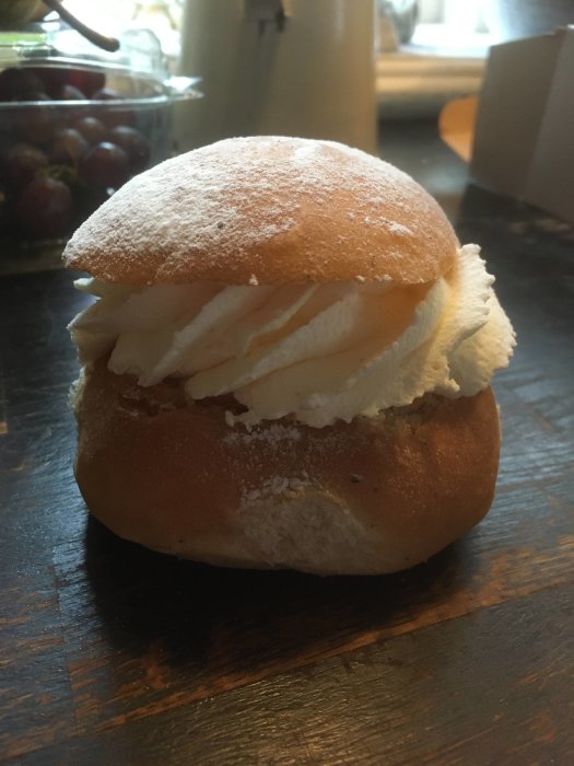 Semla med vispad grädde och florsocker, på mörkt träbord.