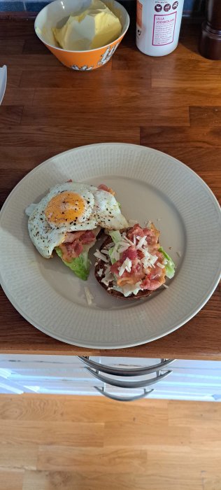 Ett fat med rågbröd, stekt ägg, bacon, sallad och riven ost. Frukost eller lunch. Träbänk och smörskål syns.