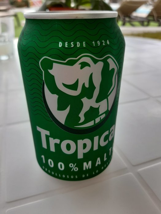 Grön burk med dryck, vågigt mönster, lejonlogotyp, text "Tropical", "100% Malta", utomhus på bord.