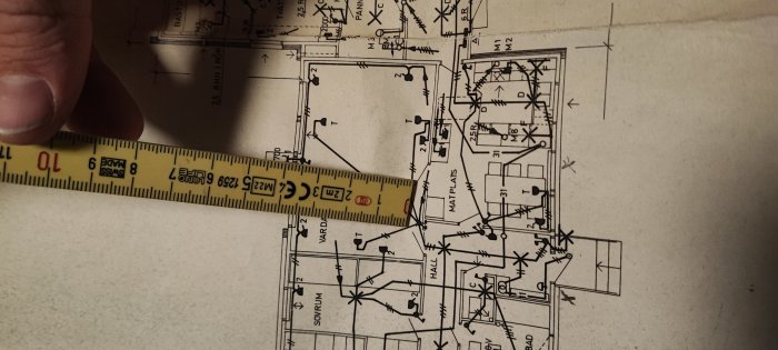 Mätningsband över byggritning, hand håller, detalj av husplan, ingenjörsmässig eller arkitektonisk kontext.