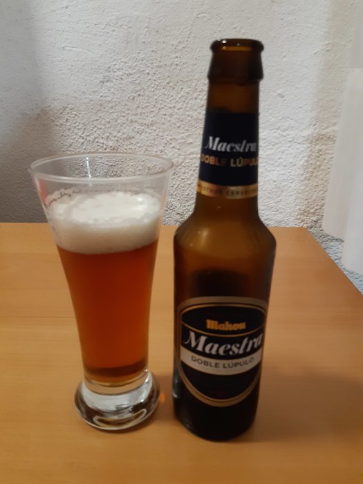 Ölglas med skum halvvägs, tom ölflaska Maestra Doble Lúpulo på bordsyta.