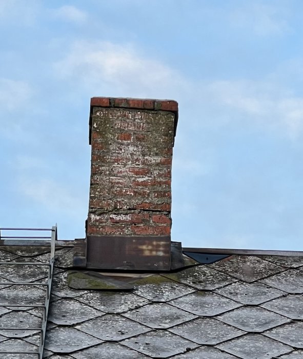 Sliten tegelskorsten på ett tak med takpannor mot en blå himmel.