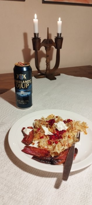 Ett bord med rösti, lingon, gräddfil, korv, en ölburk och tända ljus.