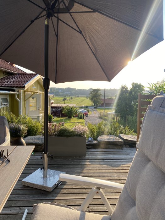 Solig veranda med parasoll, trämöbler, med utsikt över landskap och en röd stuga.