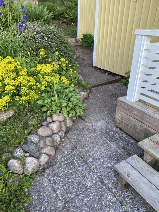 En mysig trädgårdsgång med blommor, stenkanter, trästege och gult hus.