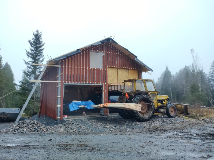 Byggarbetsplats, halvfärdigt trähus, gult fordon, grå himmel, lerig mark, byggmaterial, stödstolpar, skogsbakgrund.