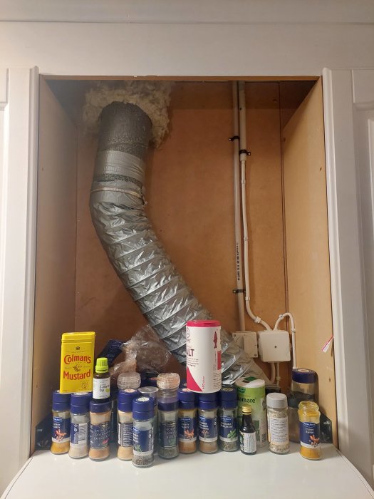 Skåp med kryddor, ventilationsslang, isolering, rör, eluttag. Oorganiserad inredning, vardagligt köksscenario.