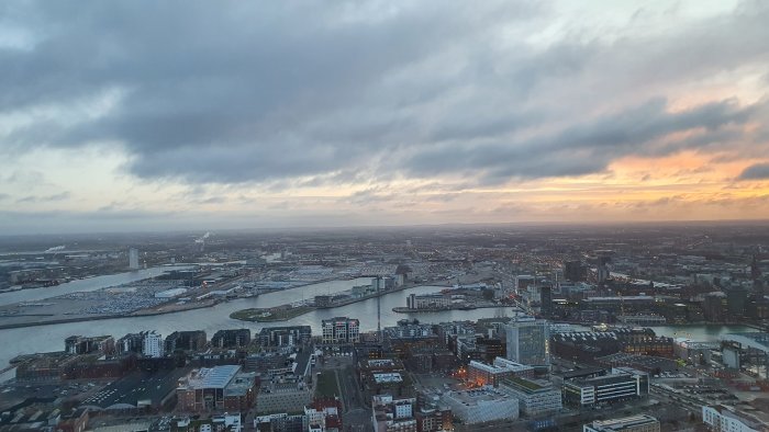 Flygfoto över stad vid solnedgång med flod, broar och byggnader. Molnigt himmel och urbant landskap.