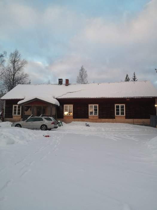Trähus täckt med snö, parkerad bil, vinterlandskap, fotspår i snön, tidig kvällshimmel.
