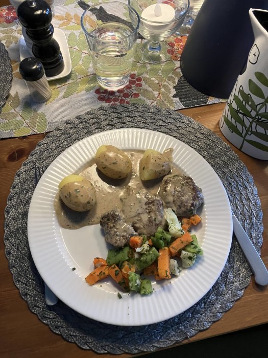 Middagstallrik med köttbullar, gräddsås, potatis och hackade grönsaker på ett bord med kryddkvarnar och vatten.