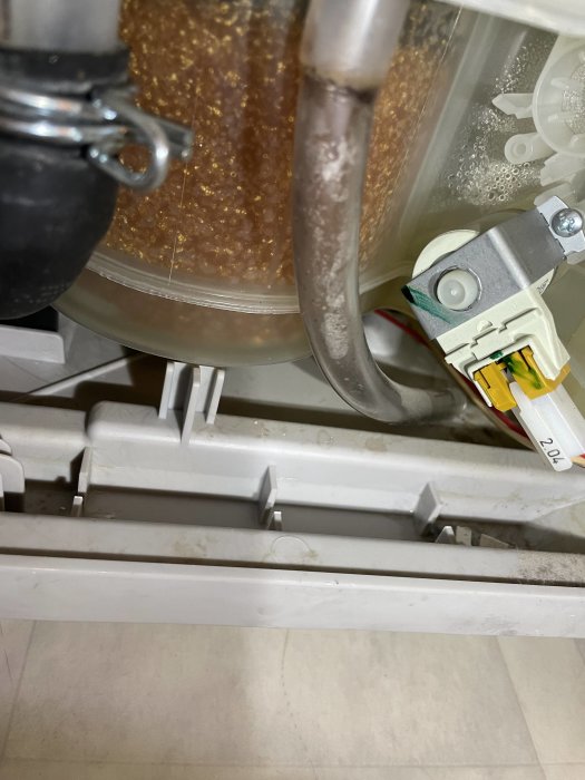 Interiör av diskmaskin med smutsiga detaljer, vattenslang, spänne och genomskinlig behållare i fokus.