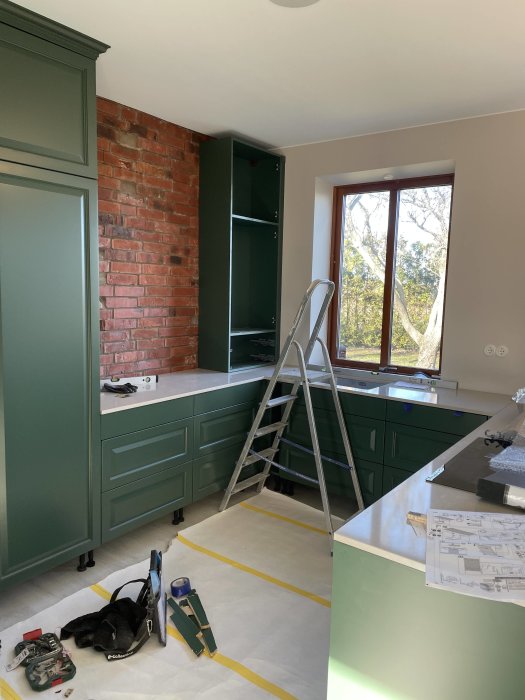 Kök under renovering med gröna skåp, rödtegelvägg, stege och verktyg på golvet.