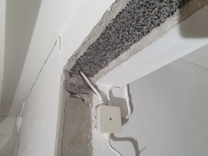 Betongvägg med skada, exponerad armering, strömkabel och vägguttag, behov av reparation.