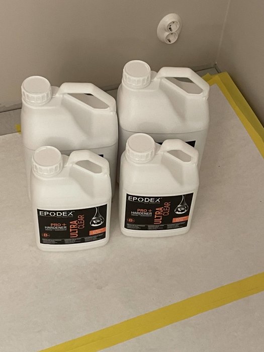 Fyra vita plastdunkar märkta "EPODEX" och "PRO+ Hardener" på ett grått golv med gul linje.