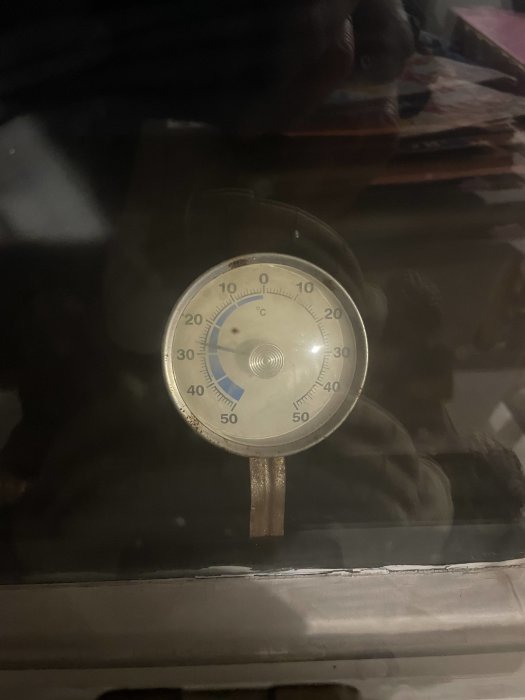 En analog termometer som visar runt 20 grader Celsius, genom en suddig, reflekterande yta.