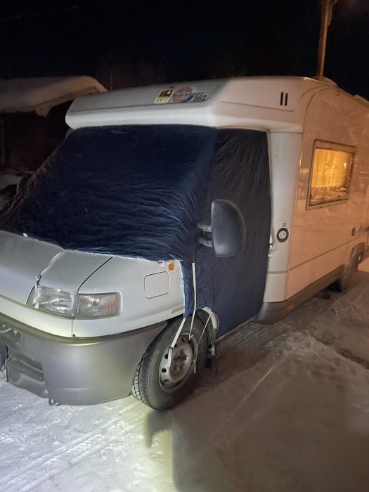 Husvagn på vintern med isolerande överdrag på vindrutan, parkerad i snö, nattbelysning, kallt väder.