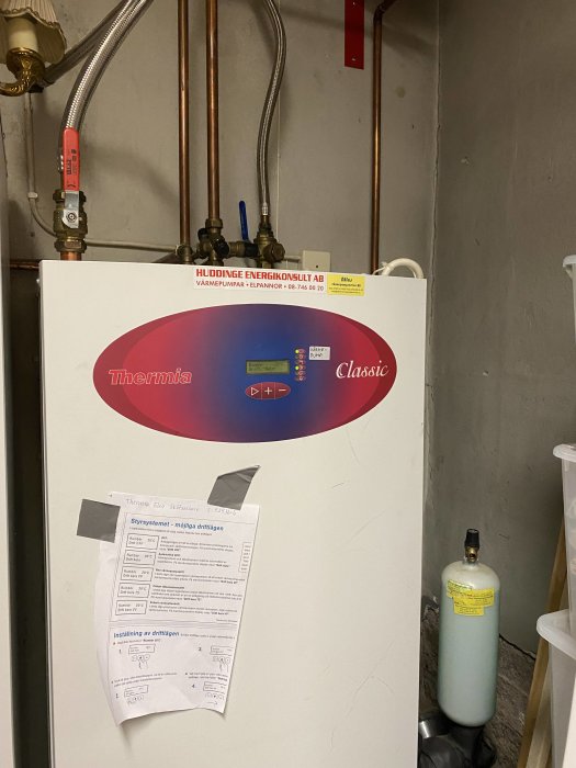 Värmepump av märket Thermia med röranslutningar, instruktionspapper, i tekniskt utrymme med expansionskärl.