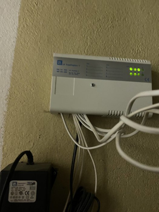 Elektrisk apparat på vägg med sladdar och lysande gröna indikatorlampor. Adapters och kablar är synliga.