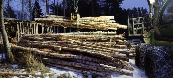 Skogsmaskin lyfter timmerstockar vid skymningen; snöigt, skogsarbete, skogsmaskineri, vinter, vedtravar.