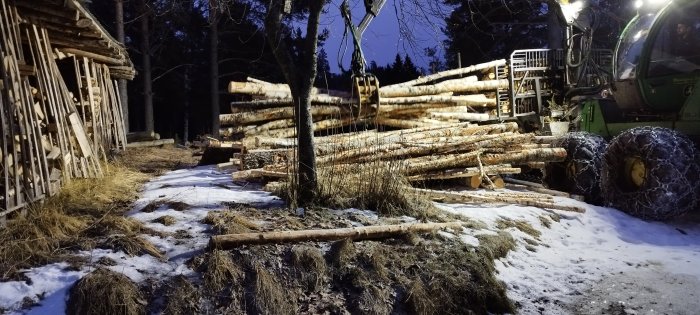 Skogsmaskin lastar timmer bredvid vedskjul i snöigt skogslandskap på kvällen.