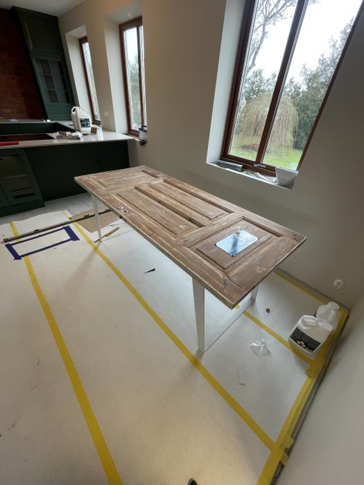 Inomhusrenovering pågår, träbord i fokus, tejpade golvlinjer, grönt köksskåp, stora fönster med utsikt, arbetsmaterial synliga.