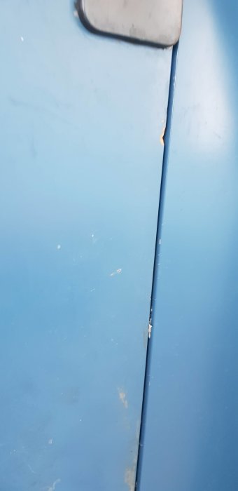 Blå yta med repor och skador, delvis täckt av grått föremål, möjlig metallstruktur eller maskindel.
