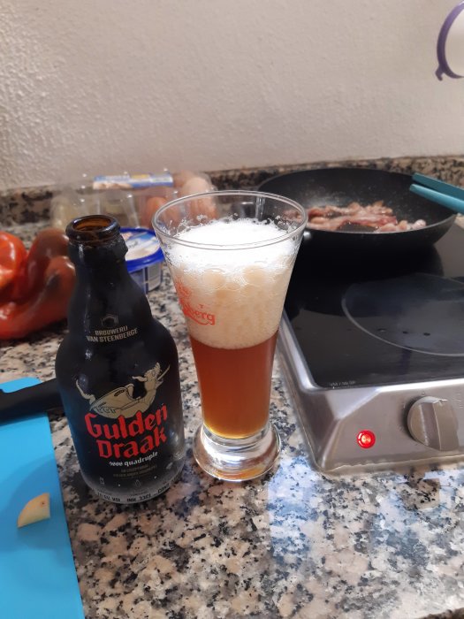 Ölflaska och glas framför spis med stekande bacon, köksbänk med matlagningsingredienser.