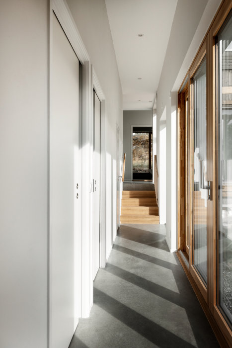 Ljus, modernt heminteriör, korridor, trätrapp, vita väggar, stora fönster, soliga skuggor, minimalistisk design.